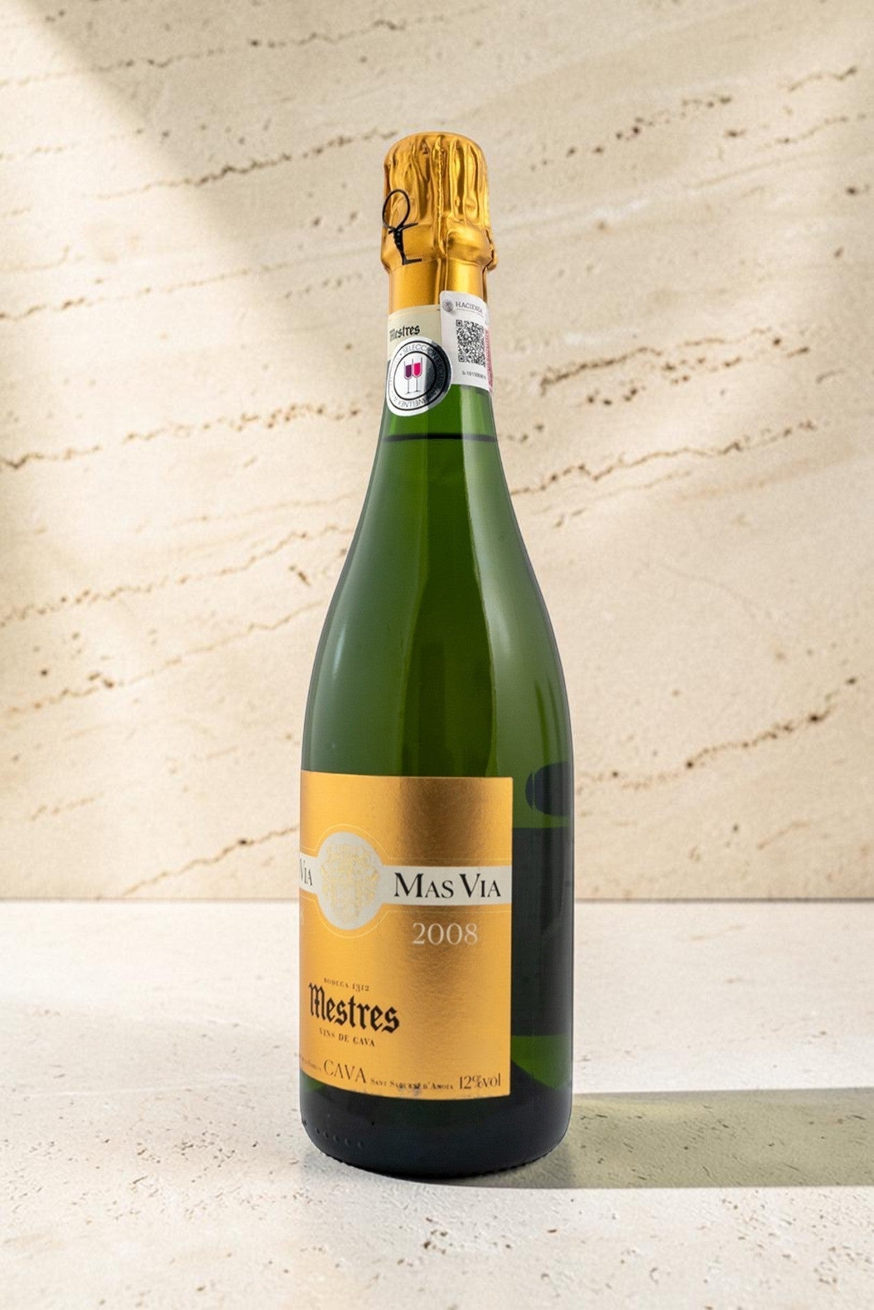 Botella de cava Mestres MAS VIA 2008 con etiqueta dorada sobre fondo de piedra texturizada en Wellnex