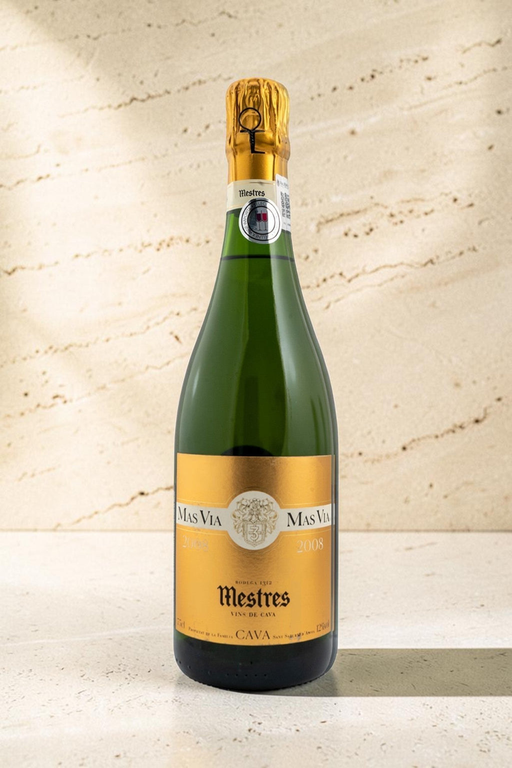Botella de cava Mestres MAS VIA con etiqueta dorada sobre fondo de piedra clara, elegante y sofisticado.