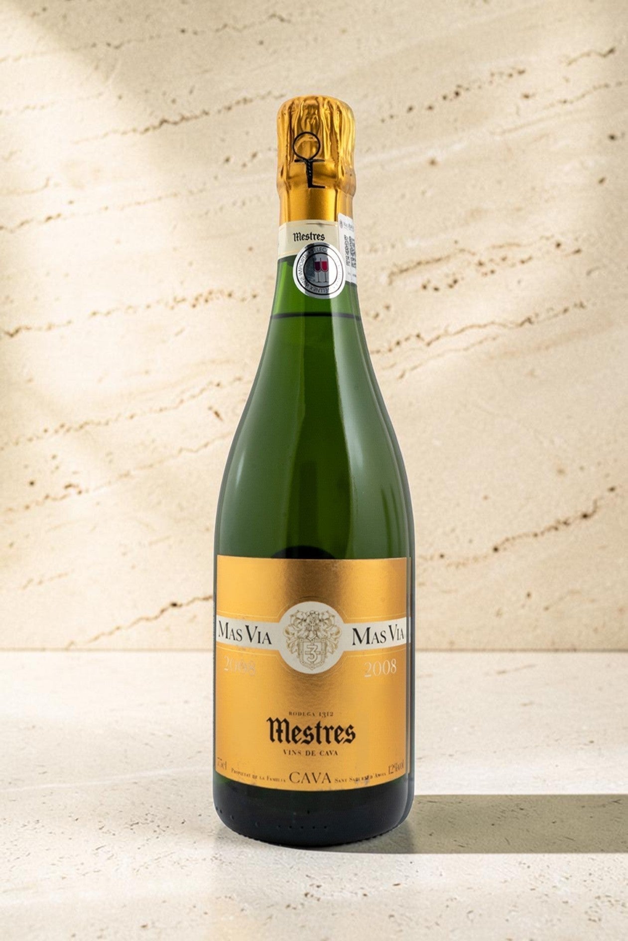 Botella de cava Mestres MAS VIA con etiqueta dorada sobre fondo de piedra clara, elegante y sofisticado.