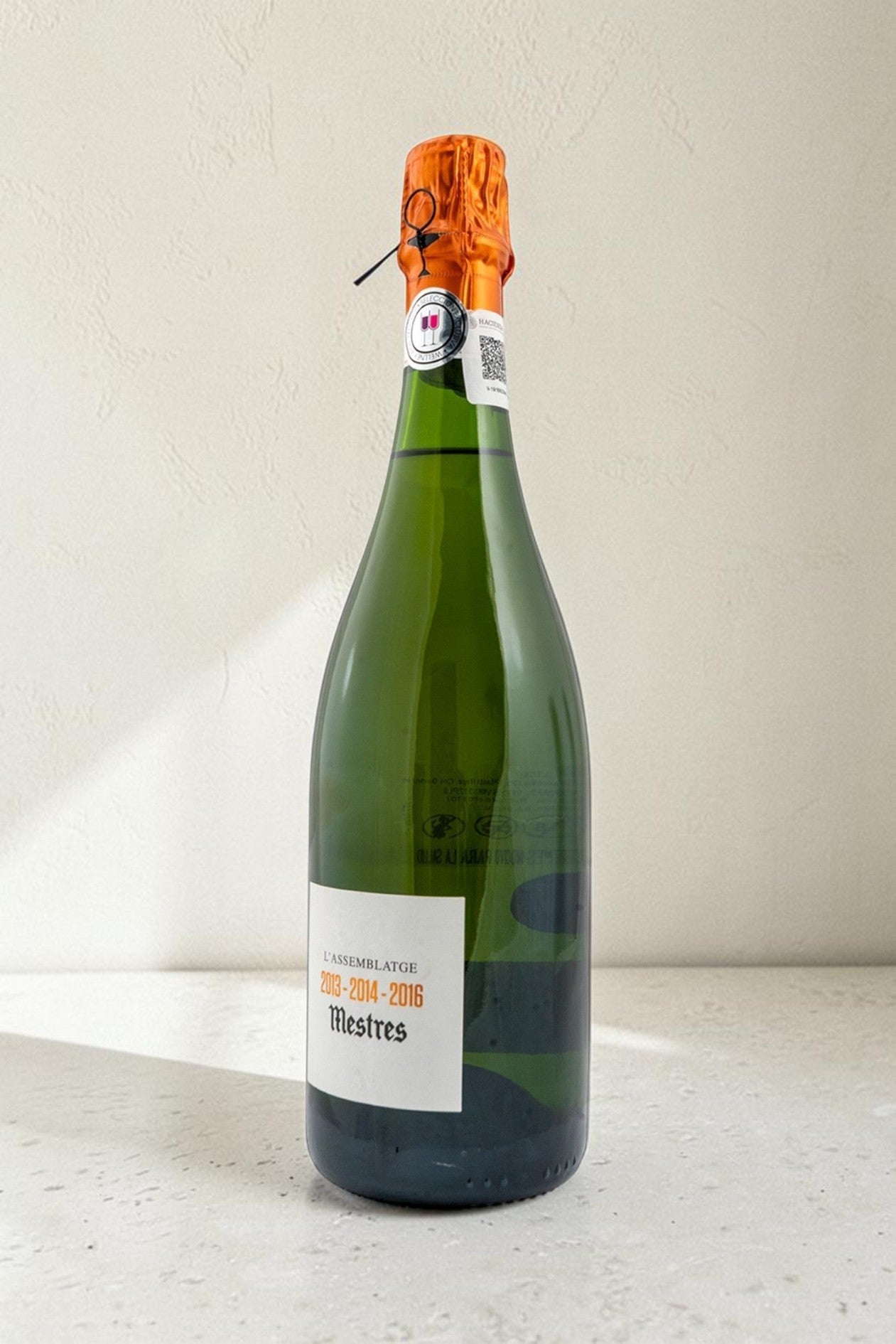 Botella de cava Wellnex Mestres Lassemblatge 13-14-16 con etiqueta blanca y cápsula naranja sobre fondo neutro