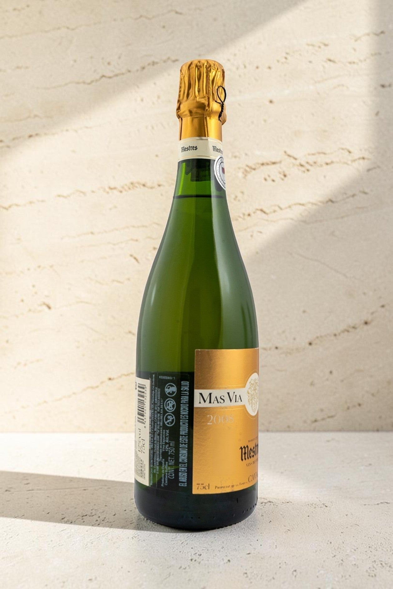 Botella de cava Wellnex Mestres MAS VIA con etiqueta dorada y cápsula brillante sobre fondo de piedra clara