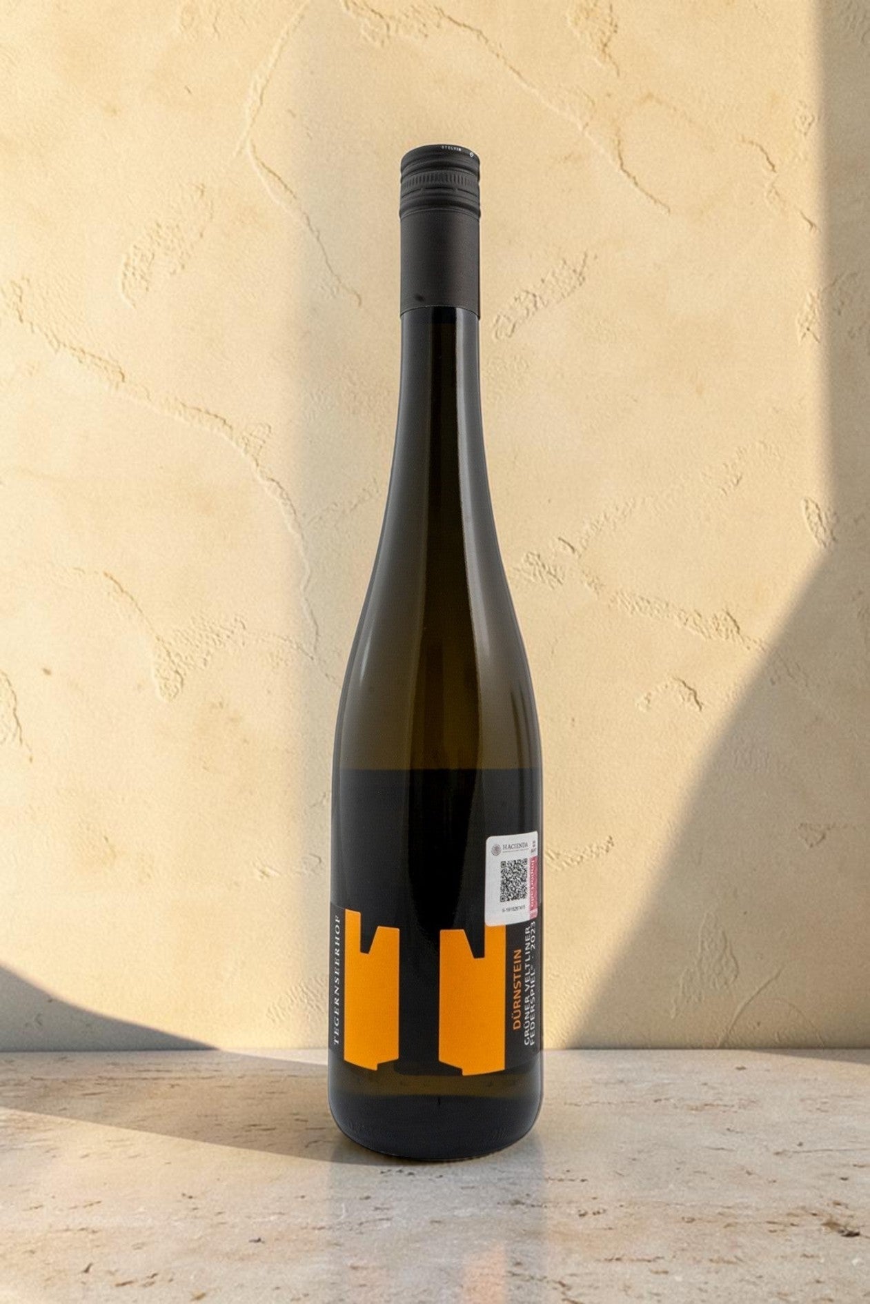 Botella de vino blanco Wellnex Weingut Tegernseerhof Gruner Veltliner Federspiel con etiqueta naranja sobre mesa.