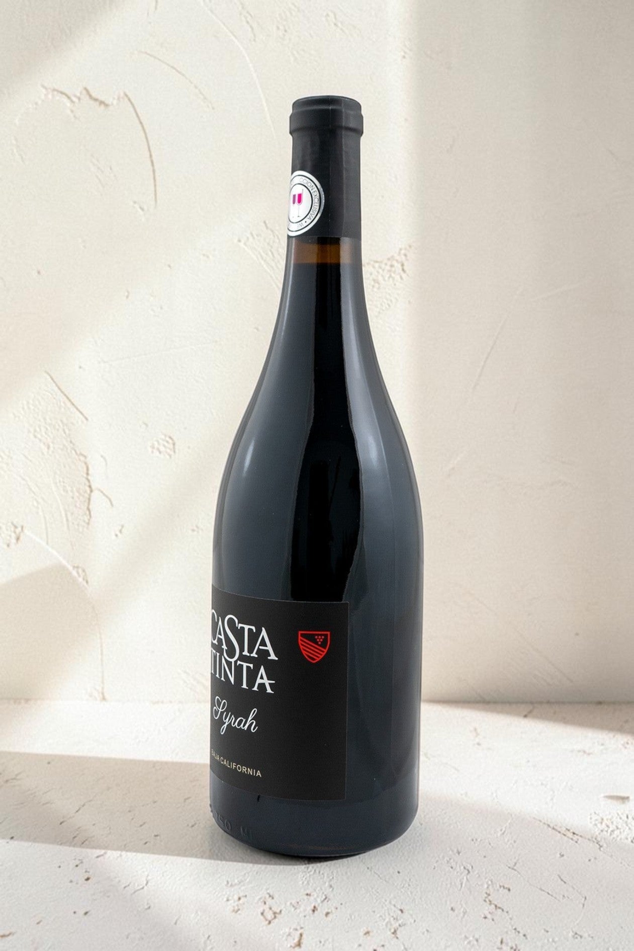 Botella de vino tinto Syrah de Wellnex con etiqueta negra sobre fondo texturizado claro