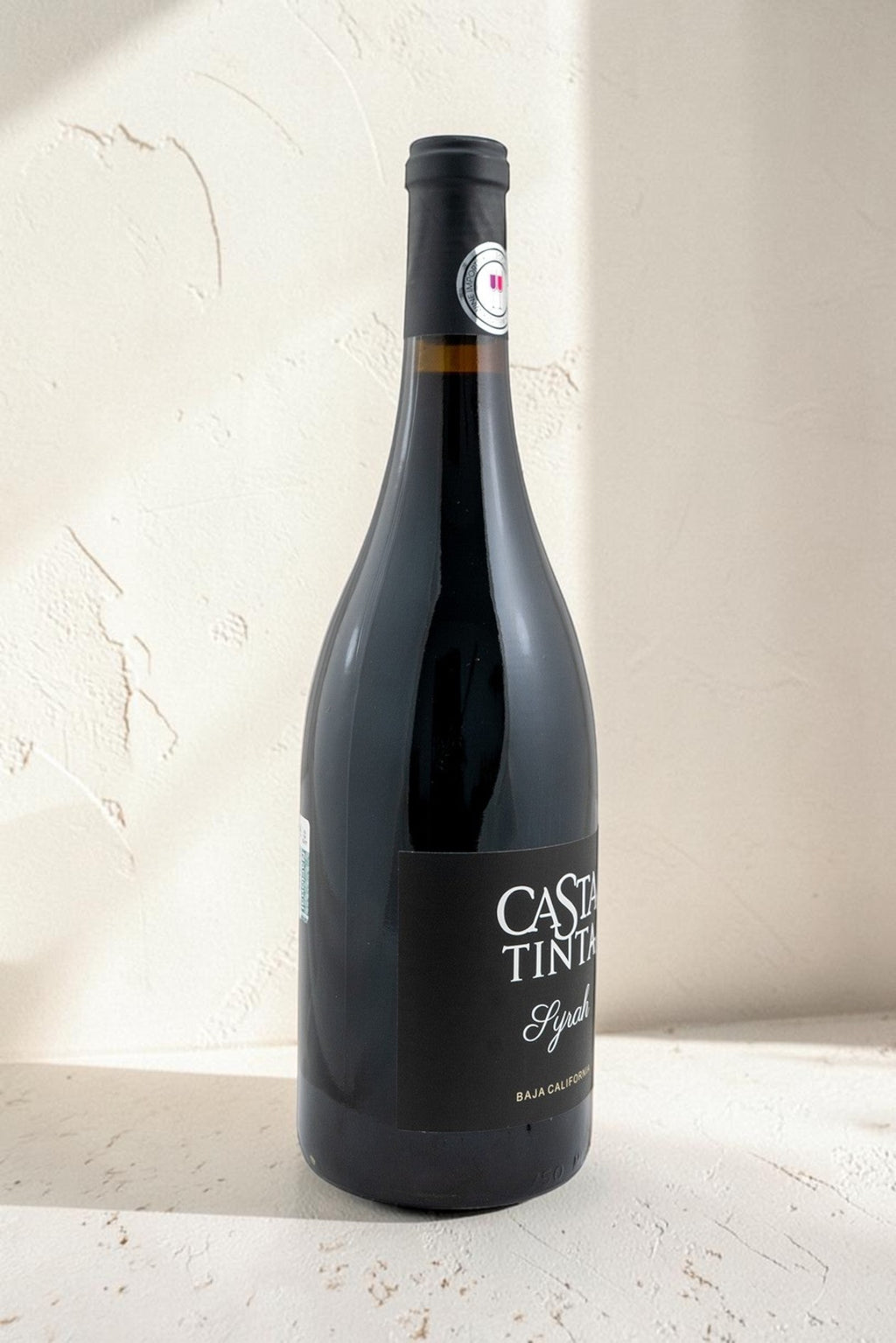 Botella de vino tinto Syrah Wellnex de color rojo rubí profundo con etiqueta elegante sobre fondo texturizado claro.