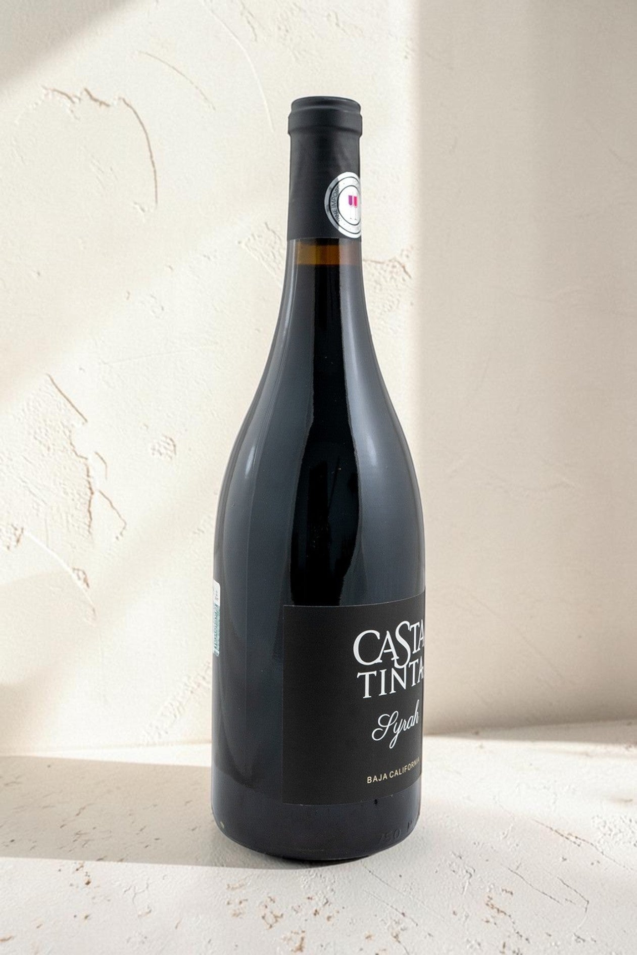 Botella de vino tinto Syrah Wellnex de color rojo rubí profundo con etiqueta elegante sobre fondo texturizado claro.